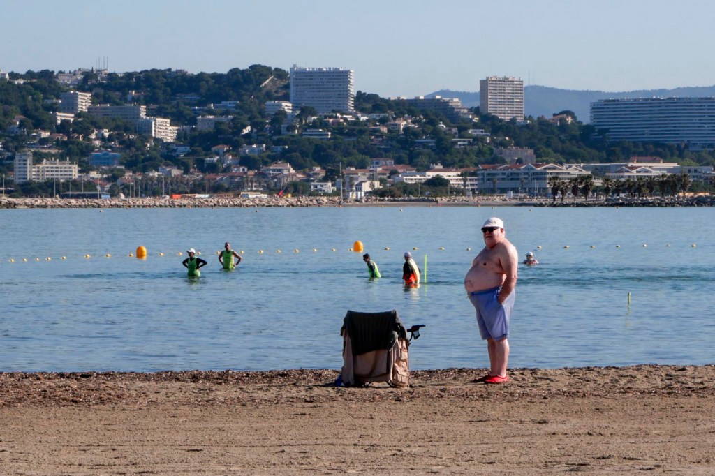 Photo couleur. Homme corpulent se tenant debout sur la plage de la Pointe Rouge, Marseille. Il tourne le dos à la plage où se trouvent des baigneurs. La baie est calme avec Marseille en arrière-plan.