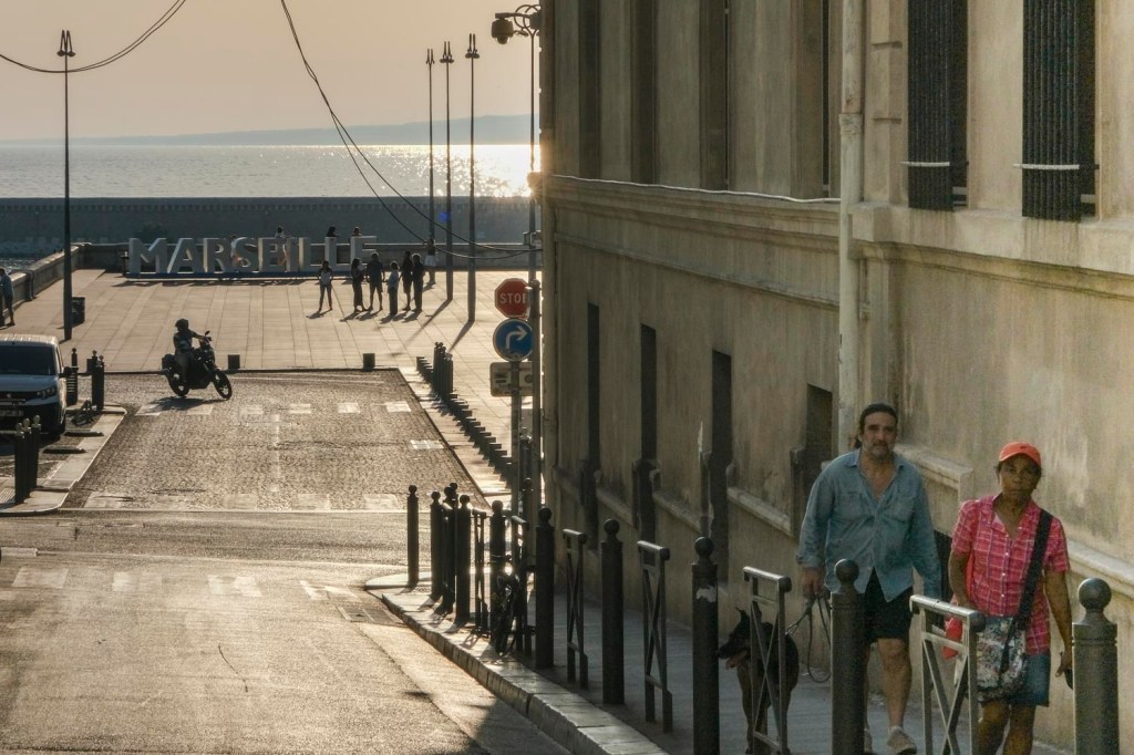 Photo couleur en contre jour. Marseille, une rue en descente arrivant sur une place où sont disposées devant la mer les grandes lettres blanches composant le nom de la ville. Petites silhouettes humaines près des lettres et la mer au fond de l'image et un couple en plan moyen à droite de l'image.
