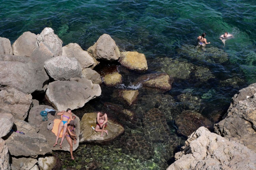 Photo couleur Marseille. Vu du dessus : la mer avec des gros rochers de couleur claire sur lesquels sont allongées ou assises trois personnes en bas à gauche de l'image tandis que dans le coin droit haut de l'image un couple se baigne dans la mer de couleur bleu vert.