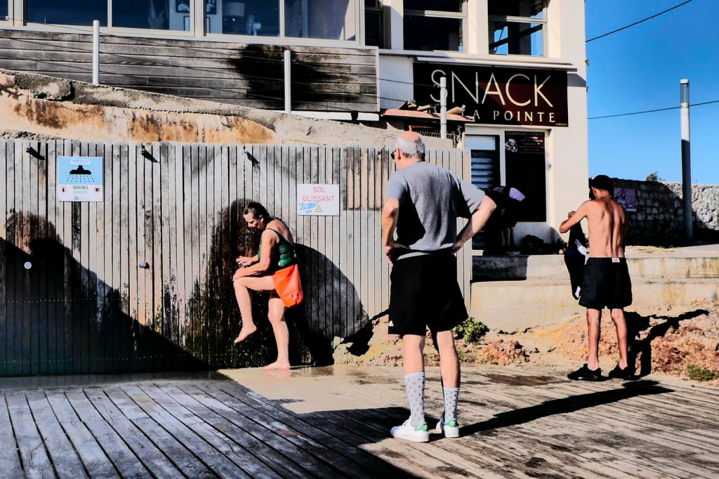 Marseille, photo couleur légèrement solarisée. Une dame sous la douche à la plage : elle fait couler de l'eau sur sa jambe sous le regard de son mari, de dos, au premier plan.
