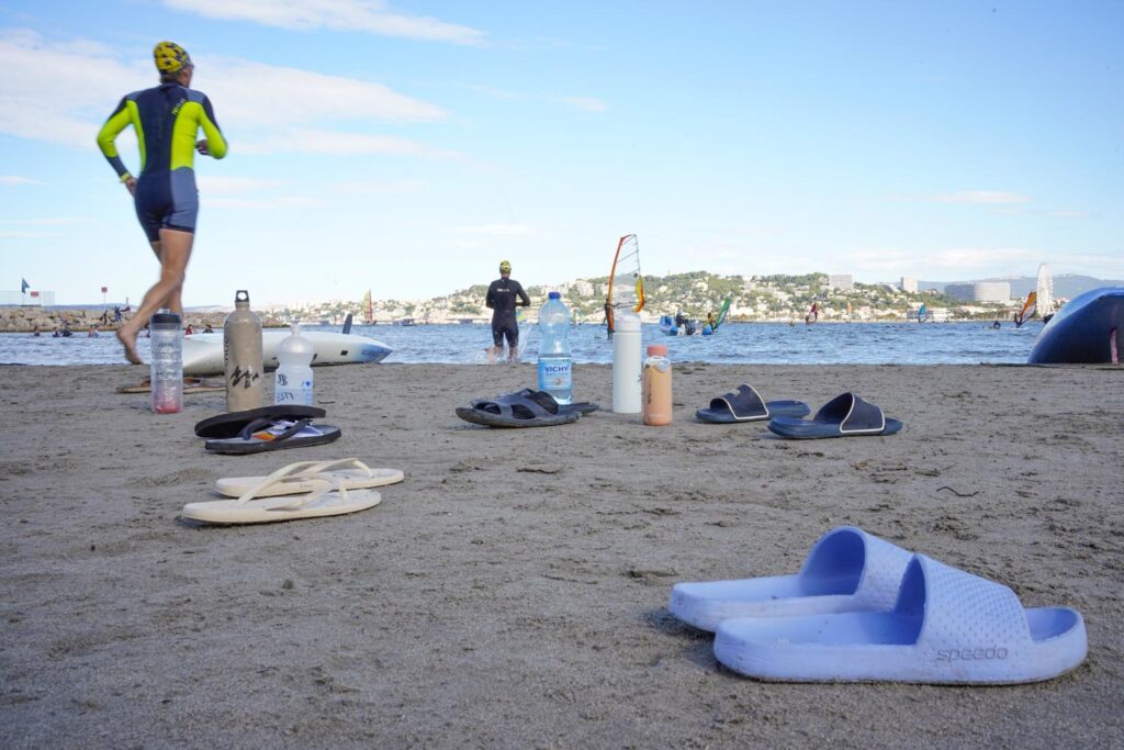 Photo couleur Marseille : la plage avec des chaussures de plage, des gourdes et deux surfeurs en tenue de surf qui courent vers la mer