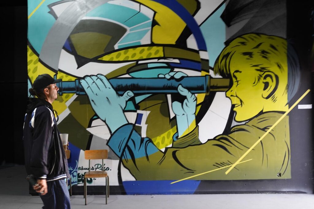 Marseille, street art. Une immense peinture d'un jeune homme de profil regardant dans une longue vue, style bande dessinée. Devant la peinture un homme arrive par la gauche, il semble avoir la longue vue dans l'œil.