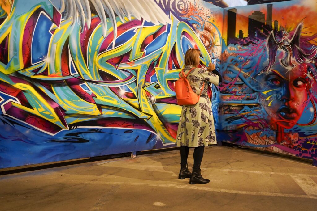 Marseille, street art. Une femme en pied de trois quart dos avec une robe verte et des collants noirs photographie une grande peinture murale très colorée.