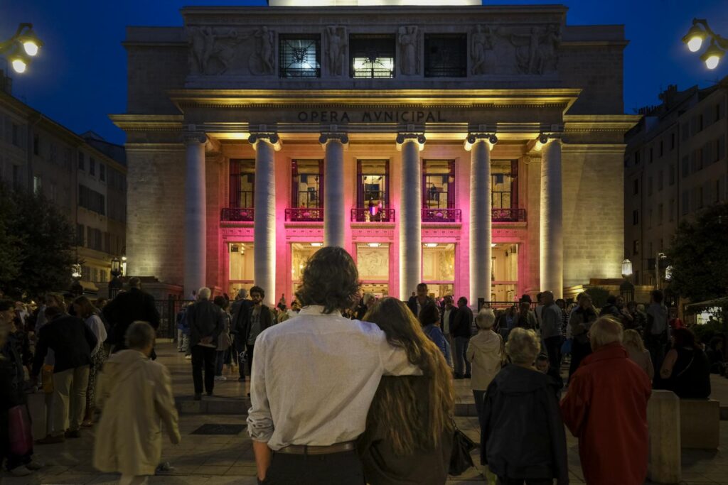 Marseille, photo couleur de nuit : la façade de l'opéra éclairée en rose. la foule sur la place devant les colonnes et un couple de dos enlacé au premier plan.